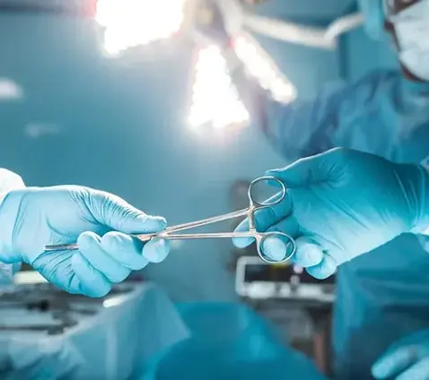 Surgeon hands surgical instrument to assistant in an operating room, both wearing gloves under bright surgical lights.