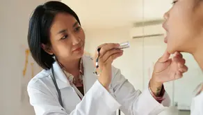 A laryngologist examines a patient’s throat with a handheld light during a clinical voice and laryngeal evaluation in medical office.