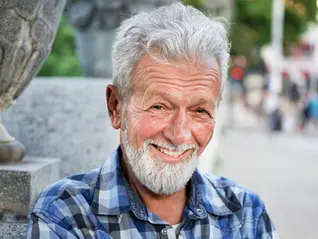 A smiling man with white hair and a white beard wears a blue and white plaid shirt. He is outdoors with a stone structure to his left and a blurred background of people and buildings.