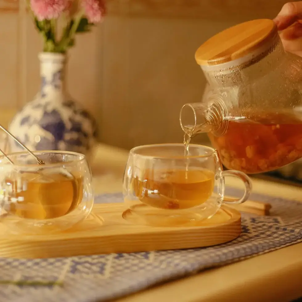 A clear glass teapot with a bamboo lid is pouring amber liquid into a clear glass teacup on a wooden tray with another teacup and a vase of pink flowers in the background.