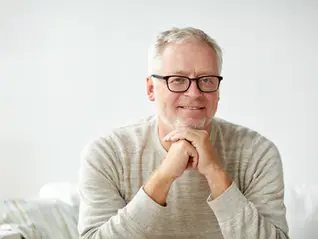 Smiling middle-aged man with gray hair and glasses rests his chin on hands while sitting on a light-colored couch.
