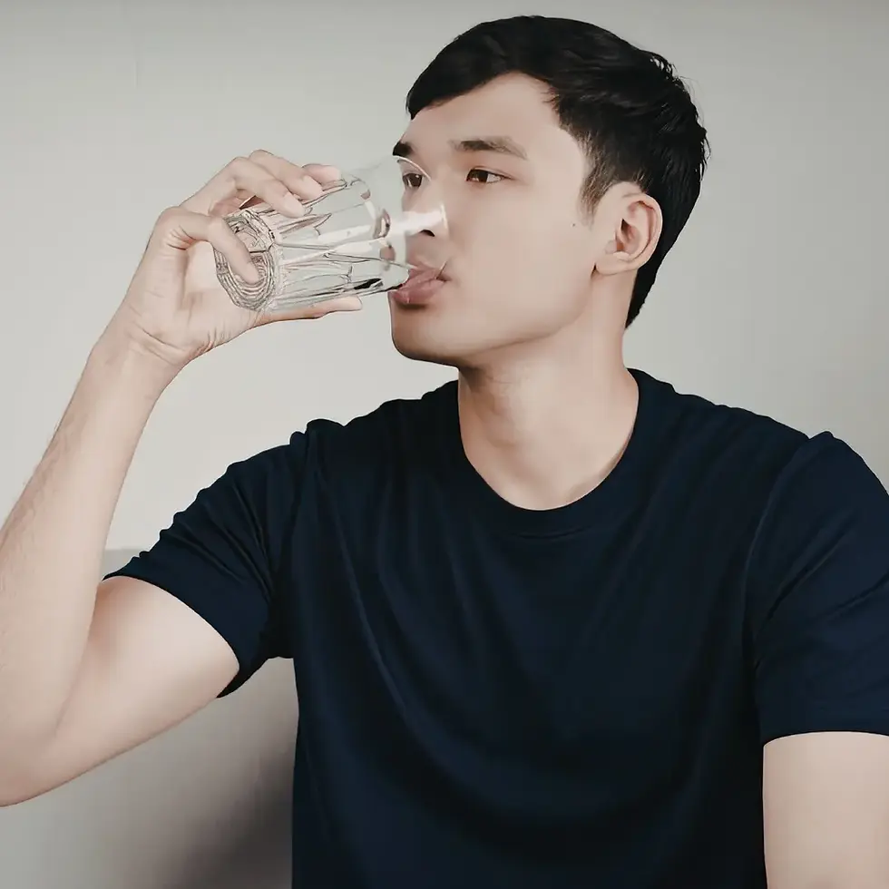 A person wearing a dark blue t-shirt drinks water from a clear glass against a plain background.
