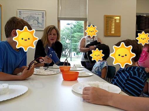 A woman at a table of kids holds a hair dryer with two paper ears and wiggle eyes.