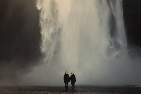 Couple in wedding attire standing at the base of Skógafoss waterfall in Iceland, dramatic and moody elopement photography.