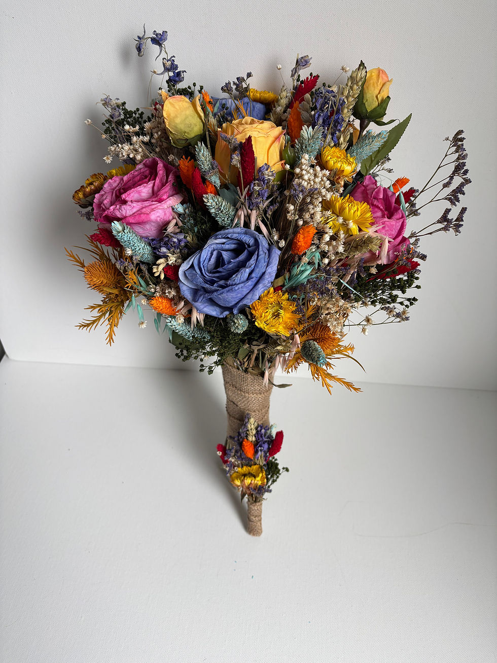 Thumbnail: Front view of a rainbow-inspired bridal bouquet and buttonhole featuring pink roses, blue roses and bright dried flowers.