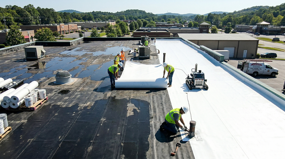 An elevated aerial view shows four workers in safety vests installing a new white single-ply membrane over a black commercial rooftop. Three workers are actively unrolling a massive sheet of material, while a fourth kneels to seal a flashing detail. Equipment including a seam welder and white buckets are visible. The background is a suburban business park with green, wooded hills.