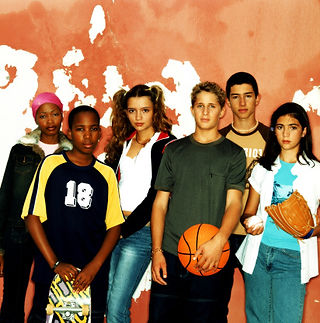 A group of young people of various genders and ethnicities stand in front of an orange wall.
