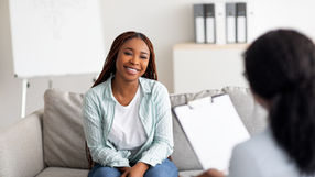 A woman sits on a couch smiling at another person holding a clip board