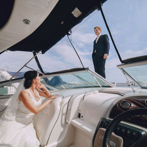 A bride admires her groom confidently standing on the yacht's deck against a backdrop of clear blue skies and a serene harbor, capturing a moment of joy and adventure on their special day.