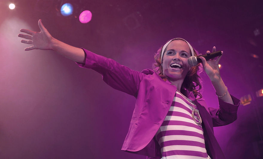 Woman singing into microphone with bright smile, in a magenta outfit