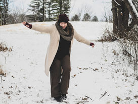 Woman in winter attire playfully walks on a snowy path in a wooded area, arms outstretched. Trees and snow-covered ground surround her.