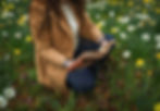 Woman in a brown coat kneels in a daffodil field, examining an old book. Green grass and yellow flowers create a serene setting.