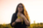 Woman with long hair holds a yellow book to her chest in a grassy field. She wears glasses, rings, and bracelets. The sky is softly lit.