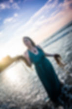 Woman in teal dress holding seaweed, standing in ocean at sunset. Waves and blue sky create a serene beach setting.