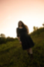 Woman in a black dress stands in a green field at sunset, with a serene expression, soft pink sky, and trees in the background.