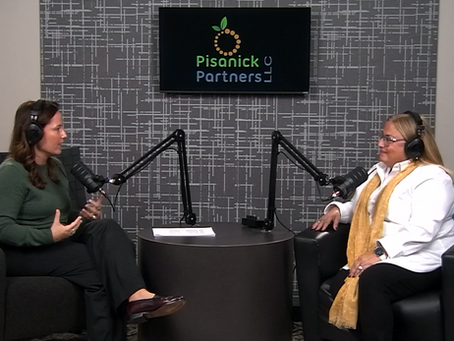 Two women wearing headsets sit across from each other in a podcast studio, speaking into microphones on boom arms at a small round table. A screen behind them displays the Pisanick Partners LLC logo, and the setting appears to be a professional podcast or interview recording.