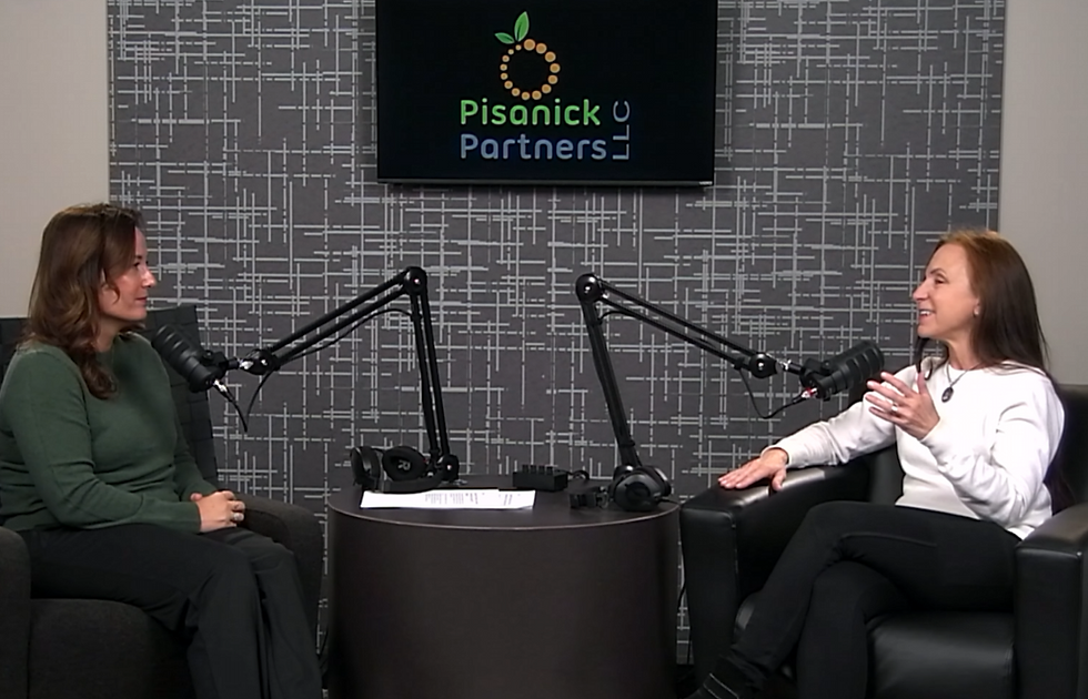 Two women talk in a podcast studio with microphones, seated under a Pisanick Partners logo on a gray patterned wall.