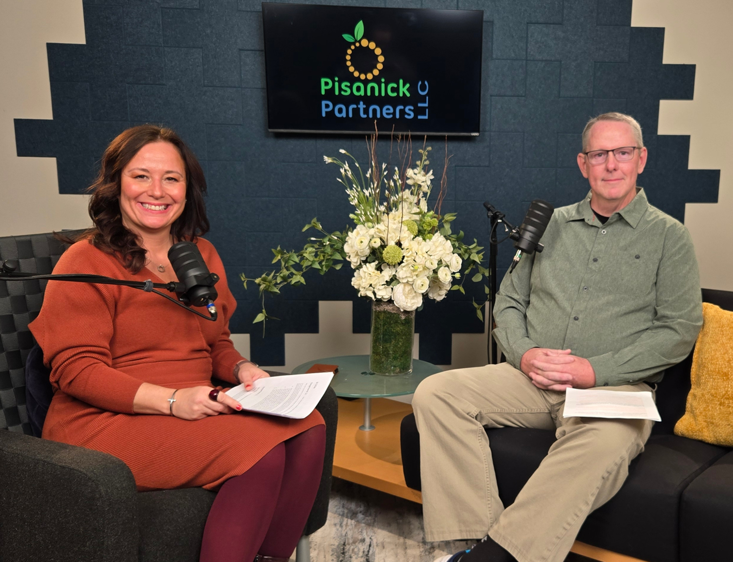 Two podcast hosts sit with microphones in a cozy studio, smiling under a Pisanick Partners LLC sign.