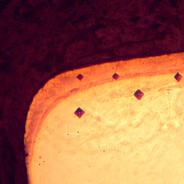 Microfotografía metalográfica de una sección transversal de acero nitrurado, mostrando cuatro huellas de indentación romboidales alineadas para medir la dureza superficial.