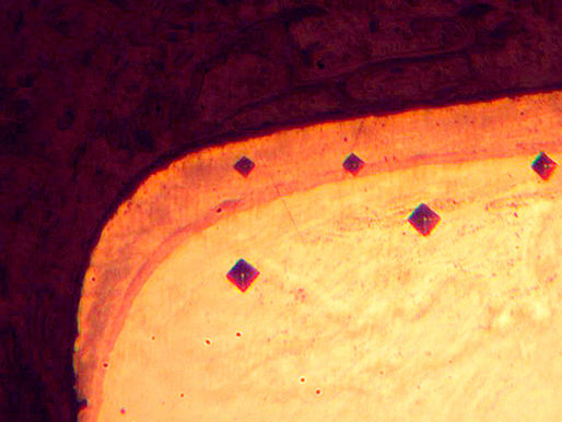 Microfotografía metalográfica de una sección transversal de acero nitrurado, mostrando cuatro huellas de indentación romboidales alineadas para medir la dureza superficial.
