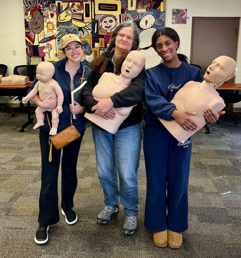 Three women are holding infant and adult CPR manikins after their first aid course in Bloomington, IN. This CPR workplace training was put together through the Sophia Travis Community Foundation grant.