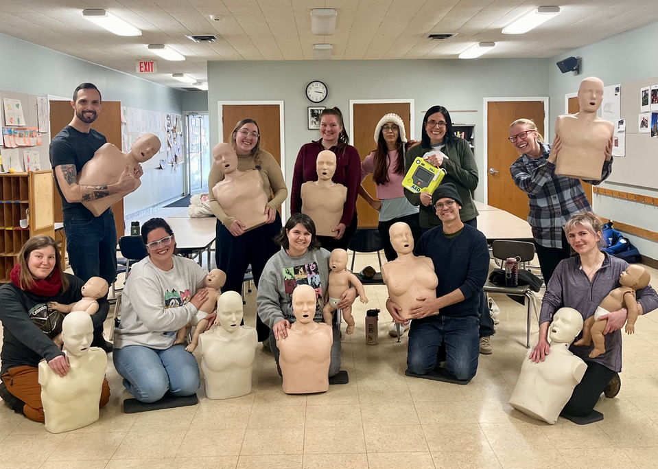 Ten teachers from the Bloomington Montessori School pose with adult and infant CPr/First Aid manikins after taking an American Heart Association First Aid/CPR training for teachers. One teacher is holding an AED.