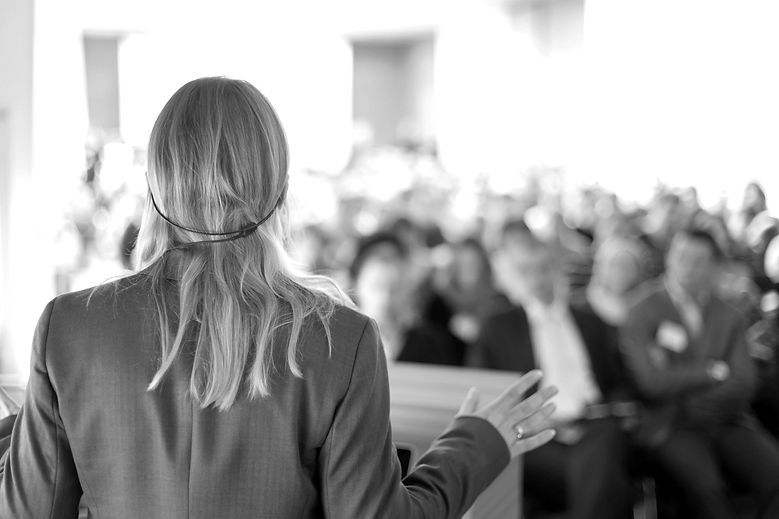 A black and white photograph taken behind a woman with long hair, wearing a headset, stood in front of a blurred crowd of people while she gives a presentation.
