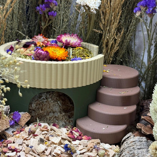 A 3D printed green circular hamster hide with an entrance at the front, brown steps at the side and cream edging along the top filled with hamster safe moss and flowers