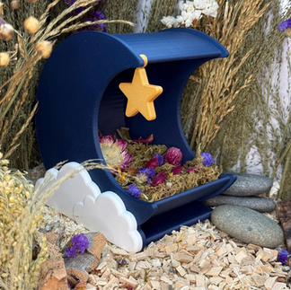 A 3D printed blue shaped moon hamster hide with a golden star hanging of the top left side and white clouds on the bottom of the side, filled with moss and flowers, surrounded by bedding, pebbles and hamster sprays
