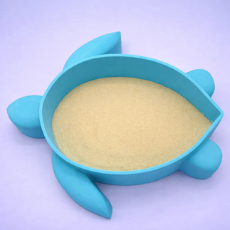 A 3D printed blue turtle shaped hamster dish filled with golden sand on a lilac purple background