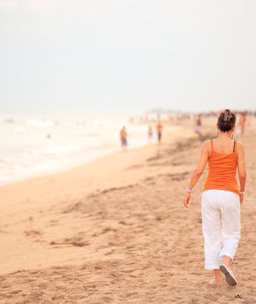Eine Frau in weißer Hose geht an einem Sandstrand entlang.