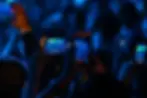 Hands hold phones recording a concert, blue lighting creating vibrant atmosphere. Stage lights visible on phone screens, capturing the live event.