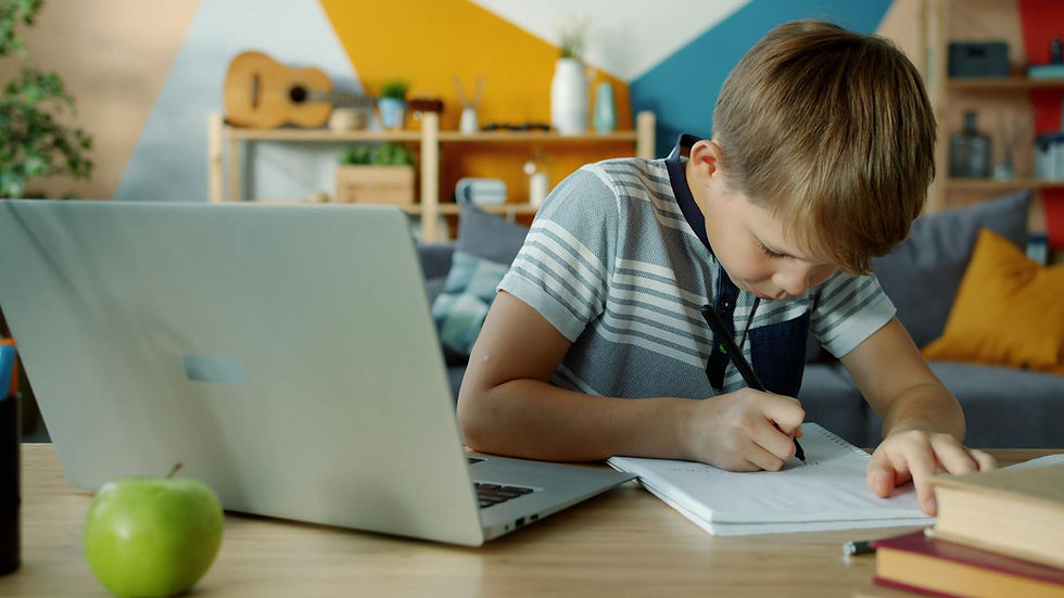 Young boy writing notes at home using a laptop for learning.