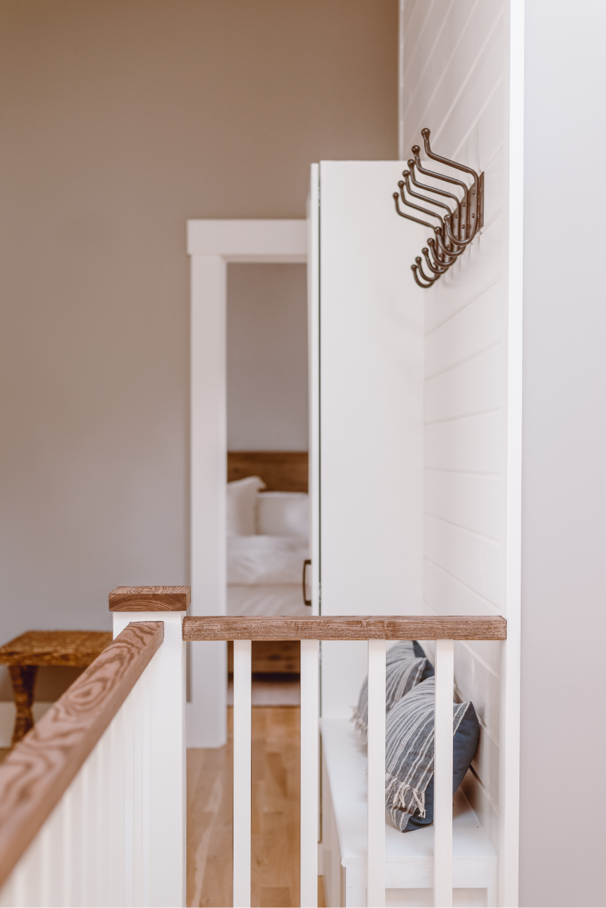 Hallway with wooden railing, hooks on a white shiplap wall, and a bench with striped cushions. A doorway leads to a bed in a softly lit room.