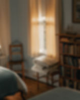 Cozy bedroom with sunlight through cream curtains, bookshelves filled with books, folded quilts on a chair, and a wooden bed frame.