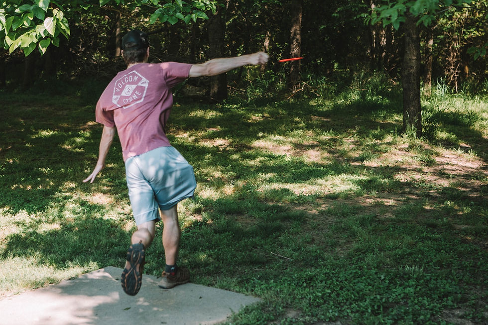 A person throws a red frisbee in a wooded park, wearing a maroon shirt and shorts. Sunlight filters through leaves, creating dappled shadows.