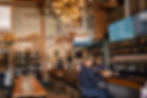 Cozy bar with rustic brick walls. A man at the counter chats with a server. Wine bottles line shelves. "WINE" sign and coffee menu visible.