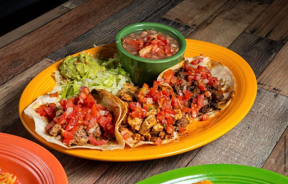 Tacos with diced tomatoes on an orange plate, alongside guacamole, lettuce, and a green bowl of salsa on a wooden table.