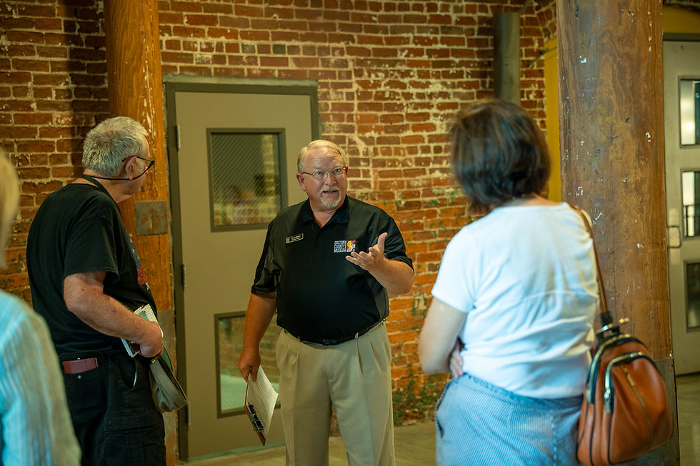 Museum guide in black polo speaks to two visitors in a brick-walled room, gesturing beside a door.