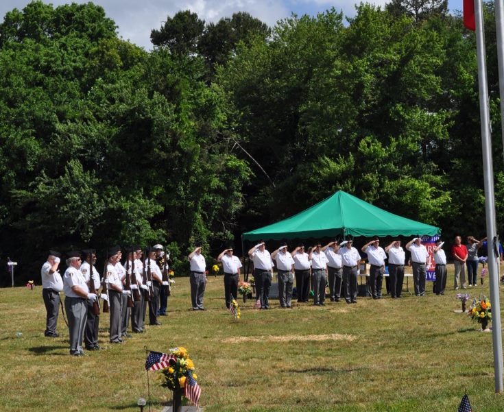 Veterans in white shirts salute in a grassy area with trees. A green tent is in the background, and small American flags are visible.
