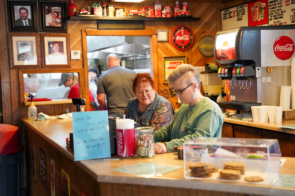 Women at a diner counter look at a menu. Background shows kitchen staff. Coca-Cola machine and signs are visible. Cozy, busy atmosphere.