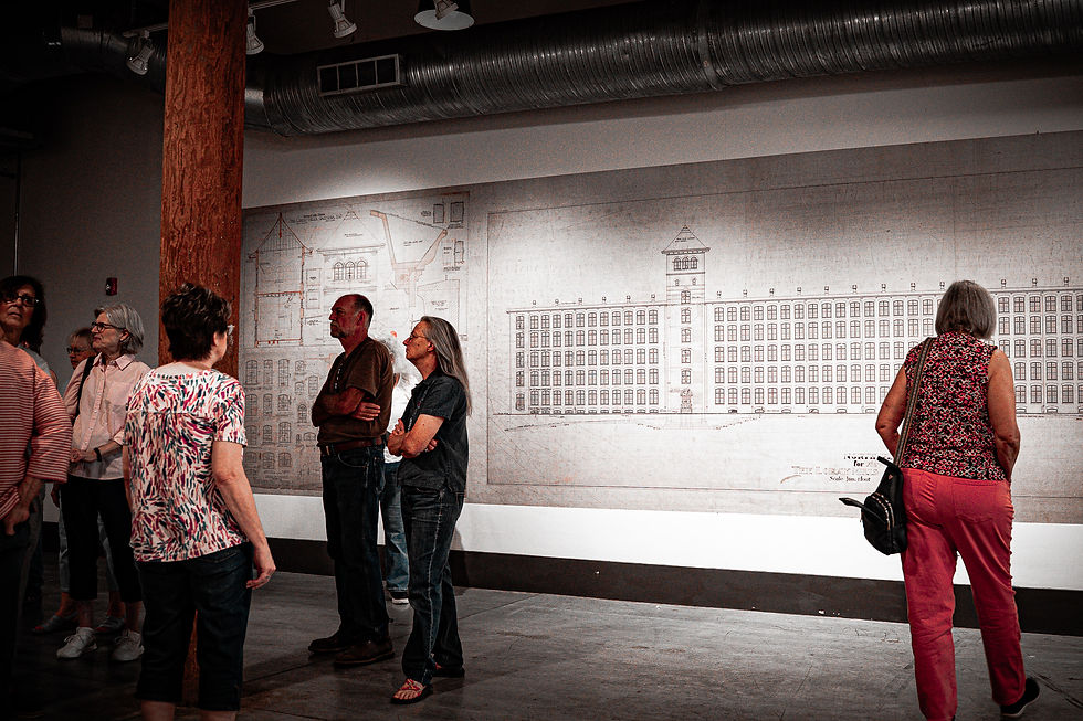Visitors stand in a gallery beside a large architectural blueprint mural in a dim industrial loft with exposed ductwork.