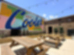 Bright patio with picnic tables and string lights beside mural reading Life is Cider Downstairs under a blue sky