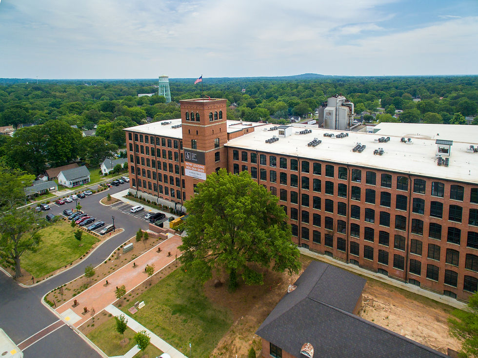 Aerial view of a brick mill building with a NOW LEASING sign, American flag, parking lot, and trees under a cloudy sky.