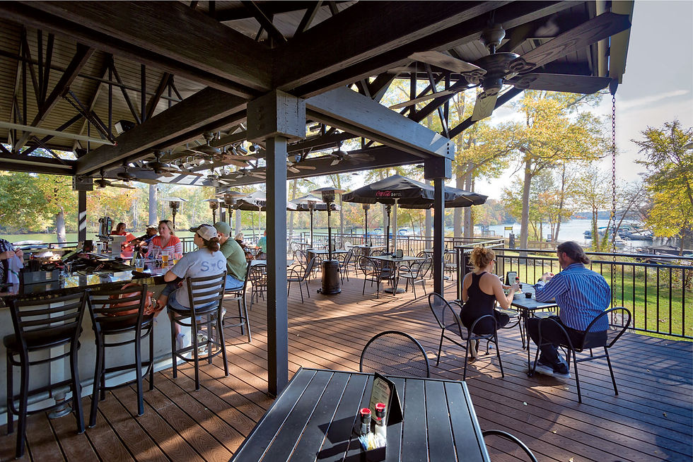 People relax on a sunny outdoor patio bar with wooden floors. Trees and a lake view in the background. Umbrellas and fans overhead.