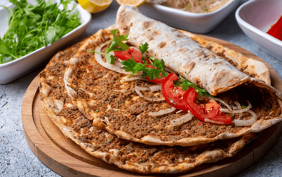 Lahmacun on a wooden board topped with sliced onions, tomatoes, and parsley. Bowls of greens and lemons are in the background.