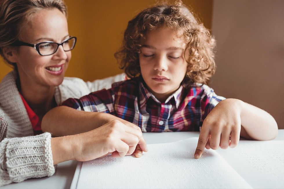 A woman guides a boy reading Braille. The boy wears a plaid shirt and focuses on the page. The woman smiles, wearing glasses and a knitted sweater.