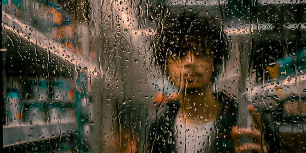 Fotografia artística do cantor de indie pop Bryan Haller vista através de um vidro embaçado e coberto por gotas de chuva. O artista aparece de forma desfocada ao fundo, criando uma atmosfera melancólica e cinematográfica