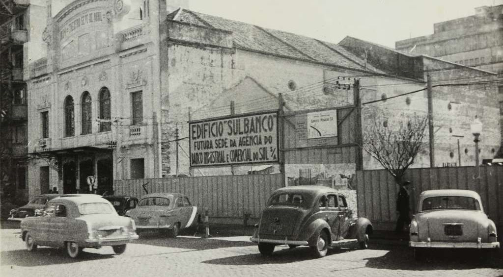 Fotografia histórica em preto e branco mostrando Edifício Sulbanco com letreiro anunciando futura sede da agência do Banco Nacional e Comercial, automóveis de época estacionados na rua de paralelepípedos, documentando transformações urbanas e comerciais em Pelotas durante período de funcionamento dos antigos cinemas mapeados pelo projeto Inventário de Plateias