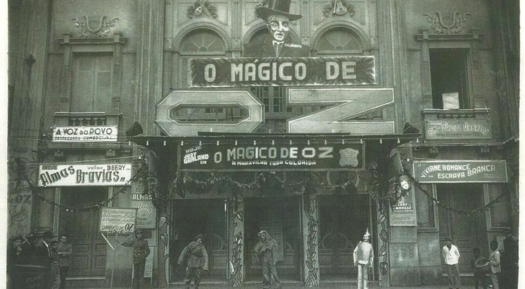 Fotografia histórica em preto e branco da fachada do Theatro Sete de Abril em Pelotas com letreiro O Mágico de Oz, decoração temática incluindo personagem com cartola no alto da fachada, pessoas posicionadas na entrada algumas possivelmente caracterizadas como personagens do filme, cartazes promocionais nas laterais, documentando as elaboradas estratégias de marketing cinematográfico utilizadas pelos cinemas pelotenses para criar experiências imersivas em torno dos filmes em exibição